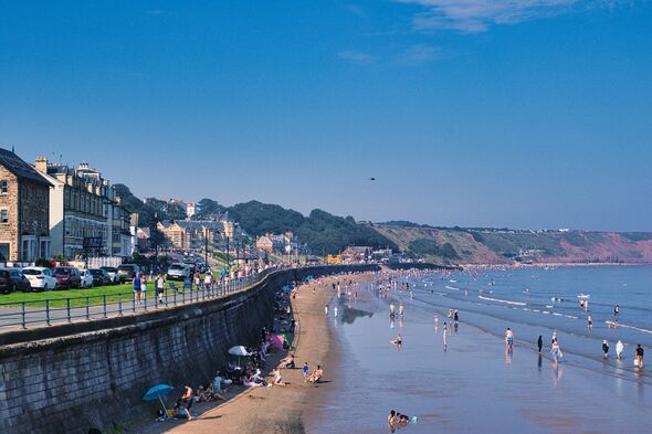 Journée de plage ensoleillée avec des gens profitant du bord de mer, du ciel bleu clair et des bâtiments côtiers en arrière-plan à Filey, en Angleterre.