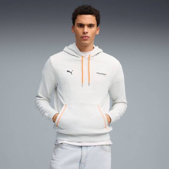 Un individu pose pour une photo tout en portant un sweat à capuche blanc avec un logo et une fermeture éclair, debout sur un dos neutre