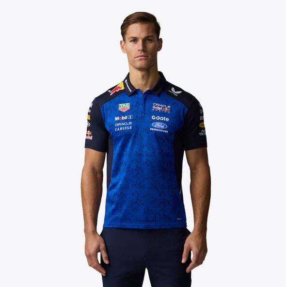 Un individu vêtu d'une chemise de sport bleue et noire avec plusieurs logos et textes de sponsors est debout et pose pour une photo.