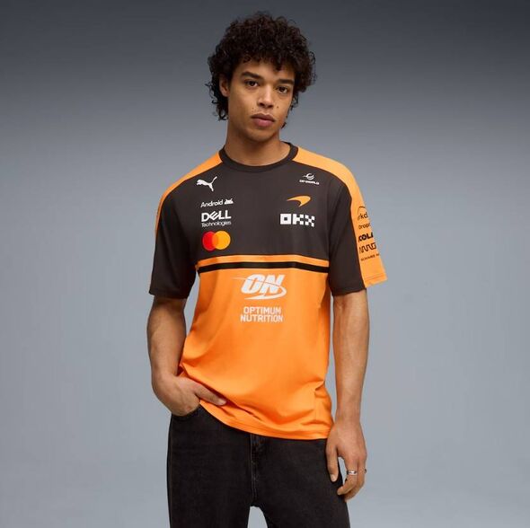 Un individu porte un maillot de sport noir et orange avec des logos de sponsors visibles, sur un fond neutre.