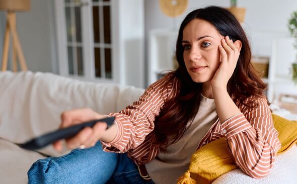 Une femme est assise sur un canapé et pointe une télécommande vers un téléviseur