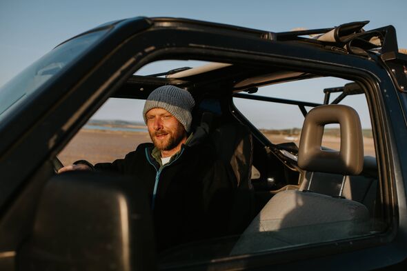 Un homme souriant portant des vêtements décontractés, dont un bonnet, conduit un véhicule tout-terrain sur une plage par une soirée ensoleillée. Il regarde